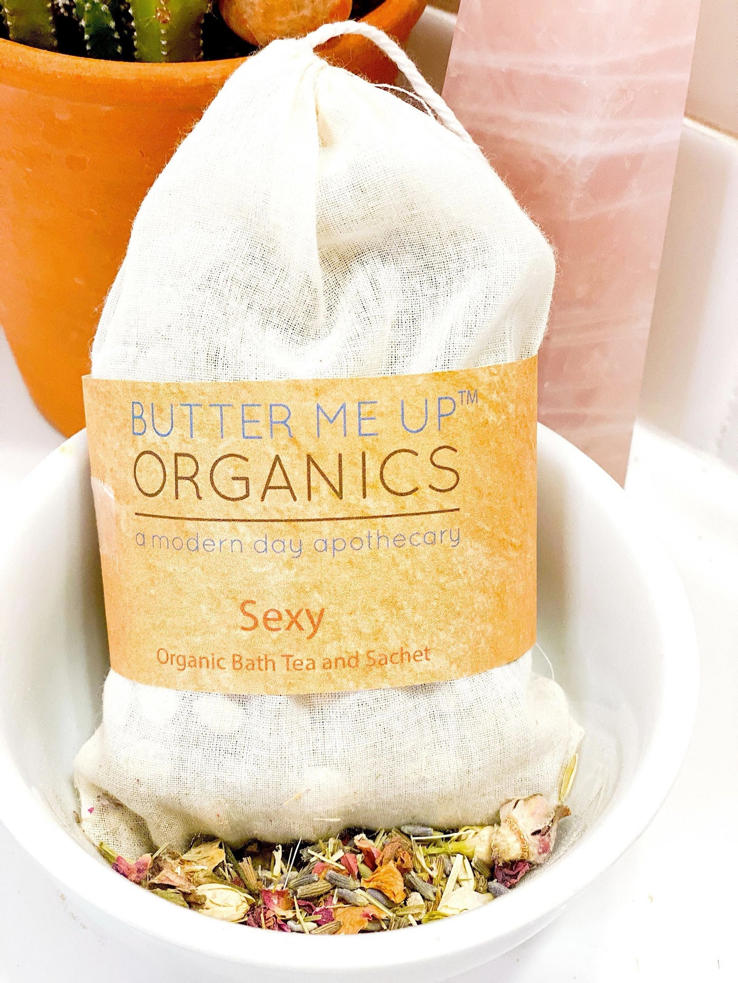In a serene setting, a cloth sachet bearing the "White Smokey: Sexy" label rests in a bowl next to loose herbs, evoking an enchanting organic bath tea experience. A potted plant and pink stone enhance the tranquility, ideal for aromatherapy.