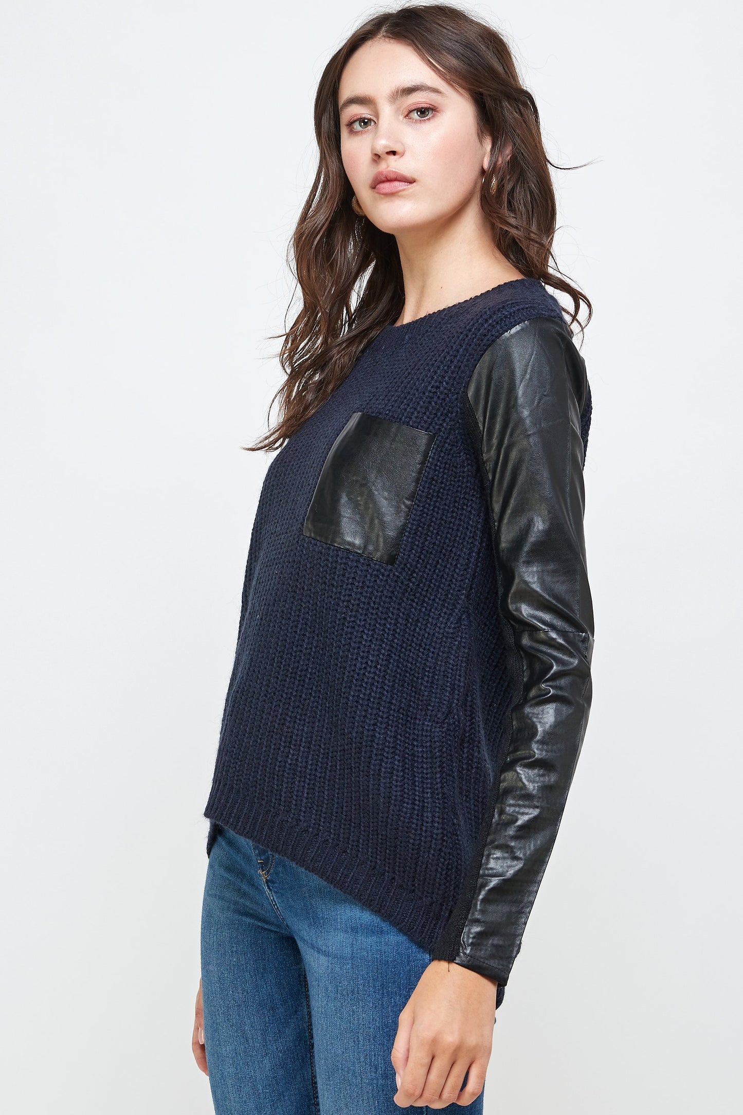 An individual models Indigo Arrowwood's Vegan Leather Sweater High Low Top with navy vegan leather, black sleeves, and a pocket, paired with blue jeans against a plain background, embodying animal-friendly fashion.