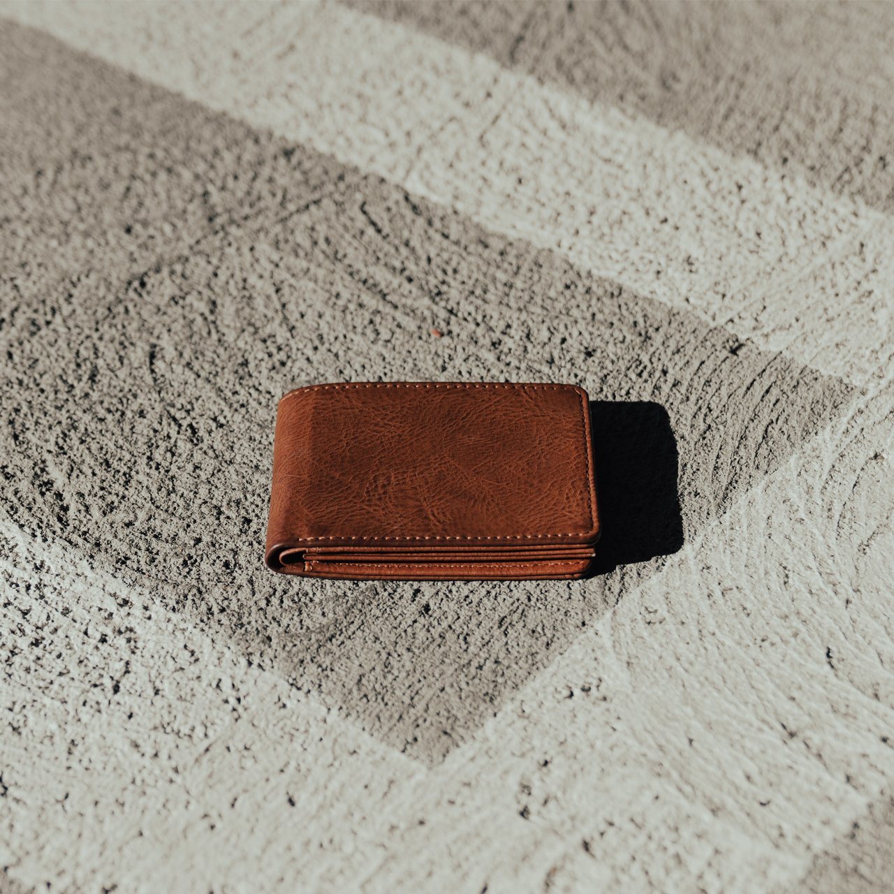 The Easy Vegan Leather Bi-Fold Wallet by Yellow Florence lies on a textured, striped concrete surface under direct sunlight, making it the perfect accessory for the conscious consumer.