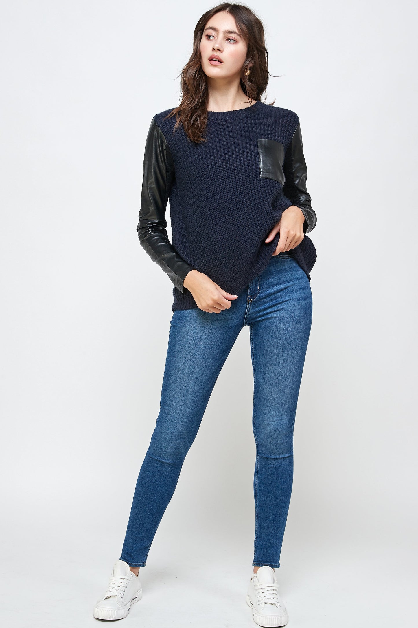 In a minimalist setting, a woman models an Indigo Arrowwood's Vegan Leather Sweater High Low Top in navy, styled with blue jeans and white sneakers, highlighting her eco-conscious fashion choice.