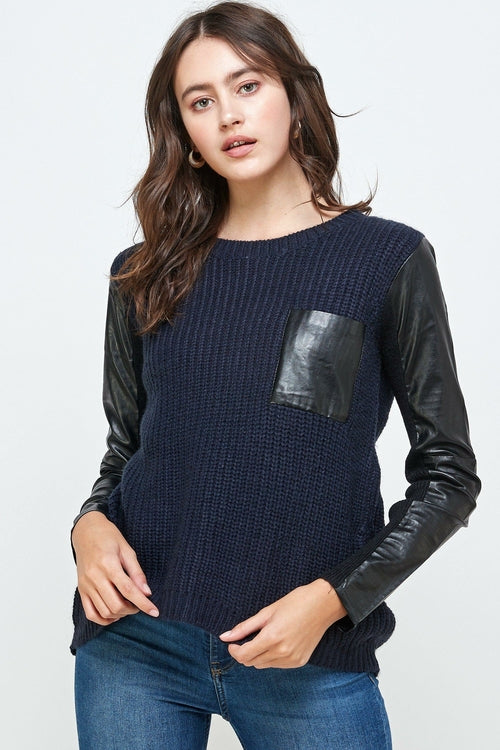 An individual dons the Vegan Leather Sweater High Low Top by Indigo Arrowwood, featuring a navy body with black leather sleeves and a pocket, embodying animal-friendly fashion against a minimalist backdrop.