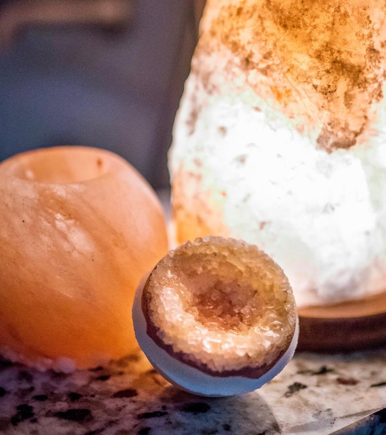 A Mint Green Icarus Himalayan Vegan Bath Bomb pairs with a glowing salt lamp and geode on a speckled surface, all harmonizing with the soothing scents of organic essential oils.