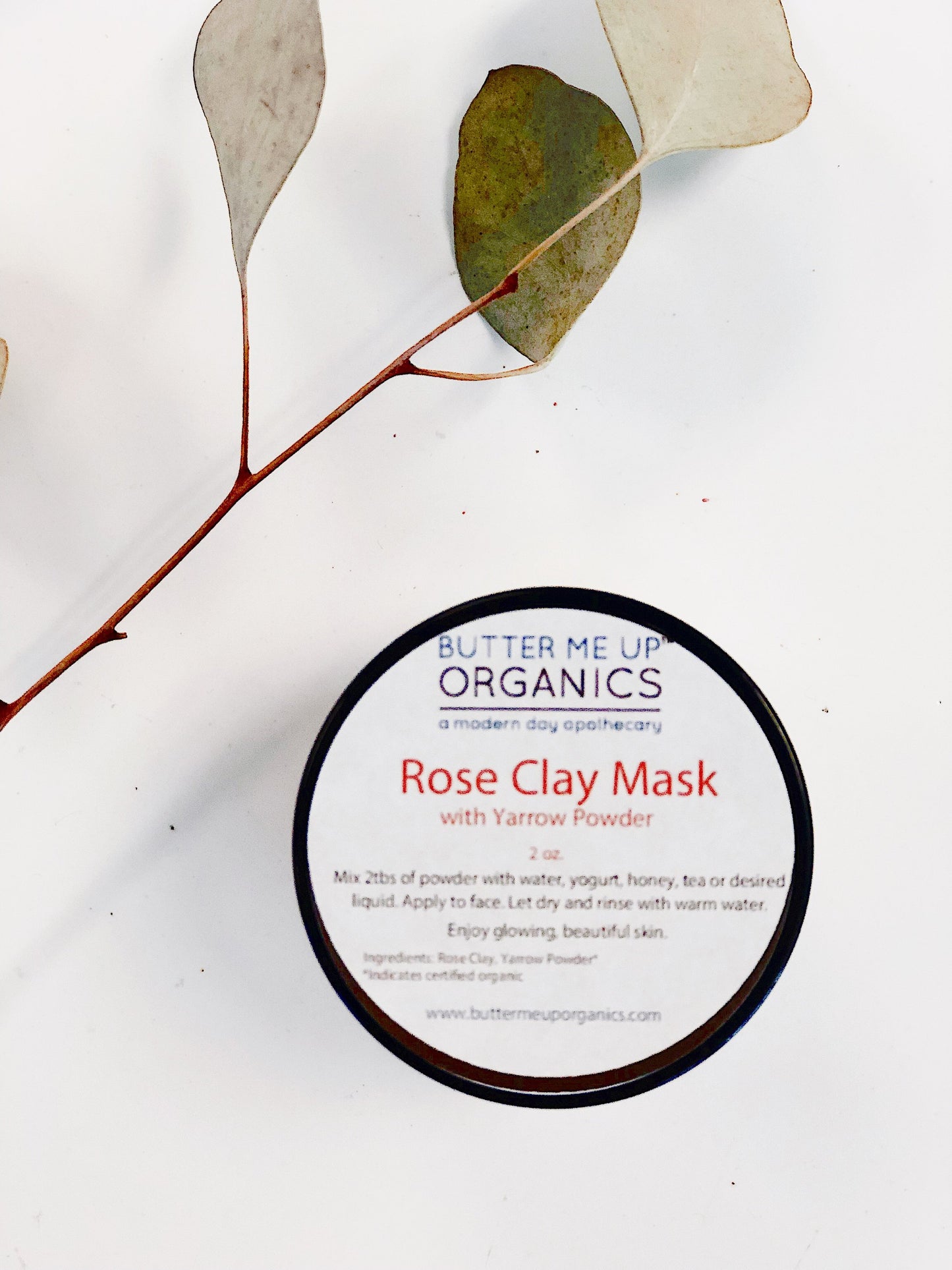 A round container labeled "Rose Clay Face Mask" by White Smokey sits on a white surface with a eucalyptus leaf beside it. Known for detoxing impurities, the mask's instructions and ingredients are listed on the lid.