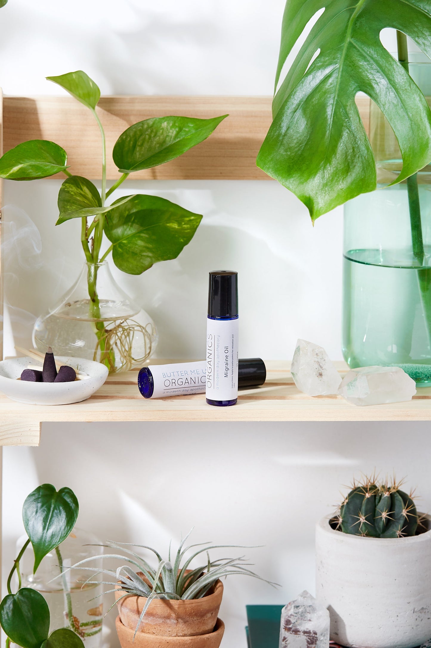A wooden shelf featuring plants, crystals, and a bottle of White Smokey's Essential Oil for Migraines provides migraine relief.