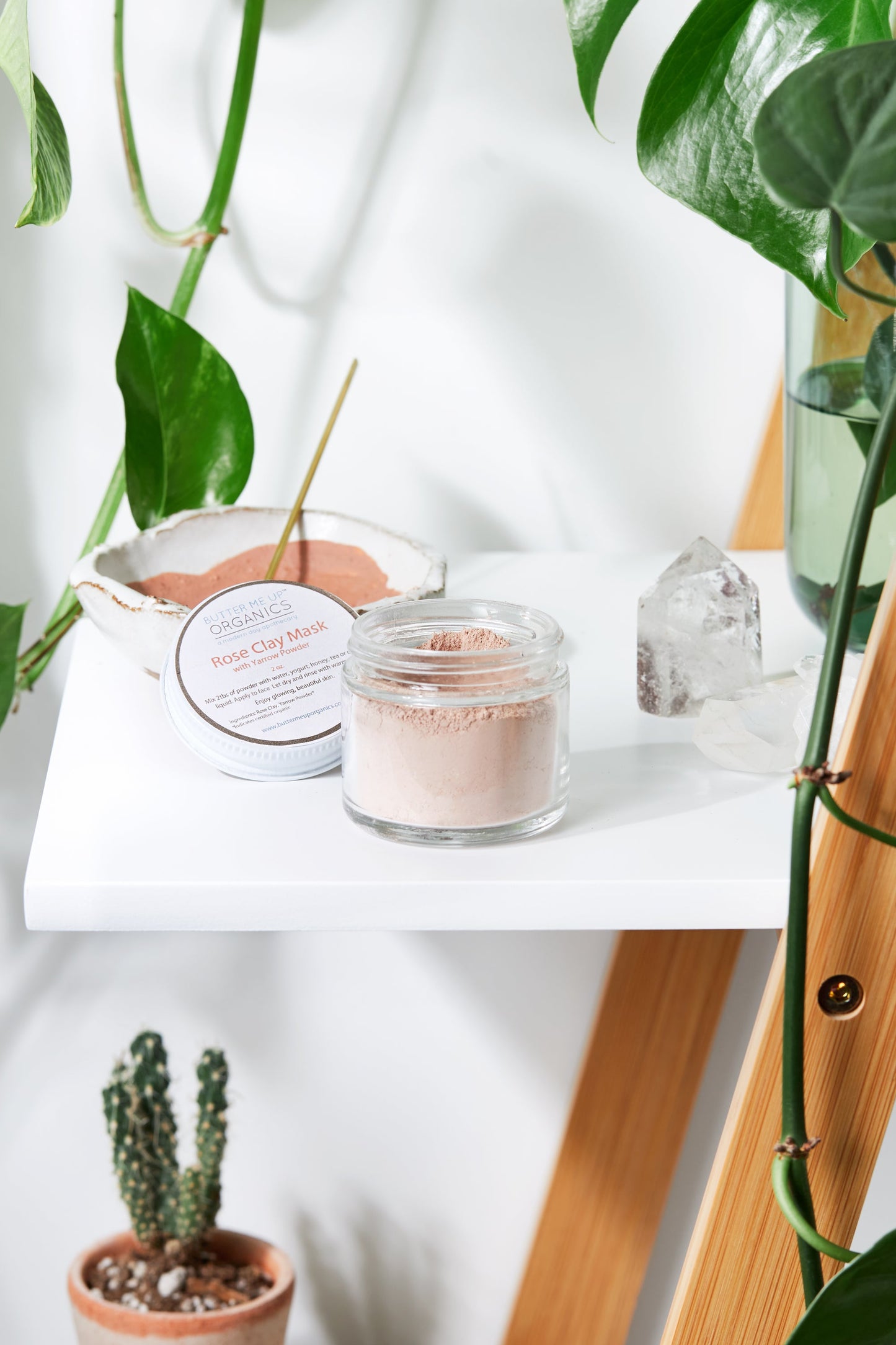 A jar of White Smokey's Rose Clay Face Mask sits on a white shelf alongside a small cactus, an incense stick, and a clear crystal. Green plants add a touch of nature both in the background and on the shelf itself.