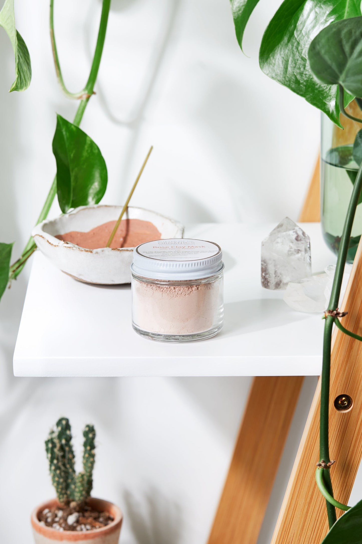 A jar of White Smokey's Rose Clay Face Mask and a ceramic bowl containing similar powder sit on a white shelf surrounded by potted plants and a crystal, perfect for detoxing impurities and improving circulation. A vertical wooden ladder is partially visible.