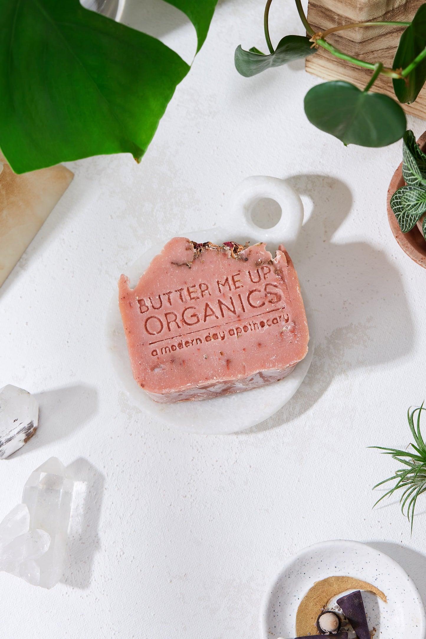 A white table with White Smokey soap and plants, including a Rose Garden Organic Soap.