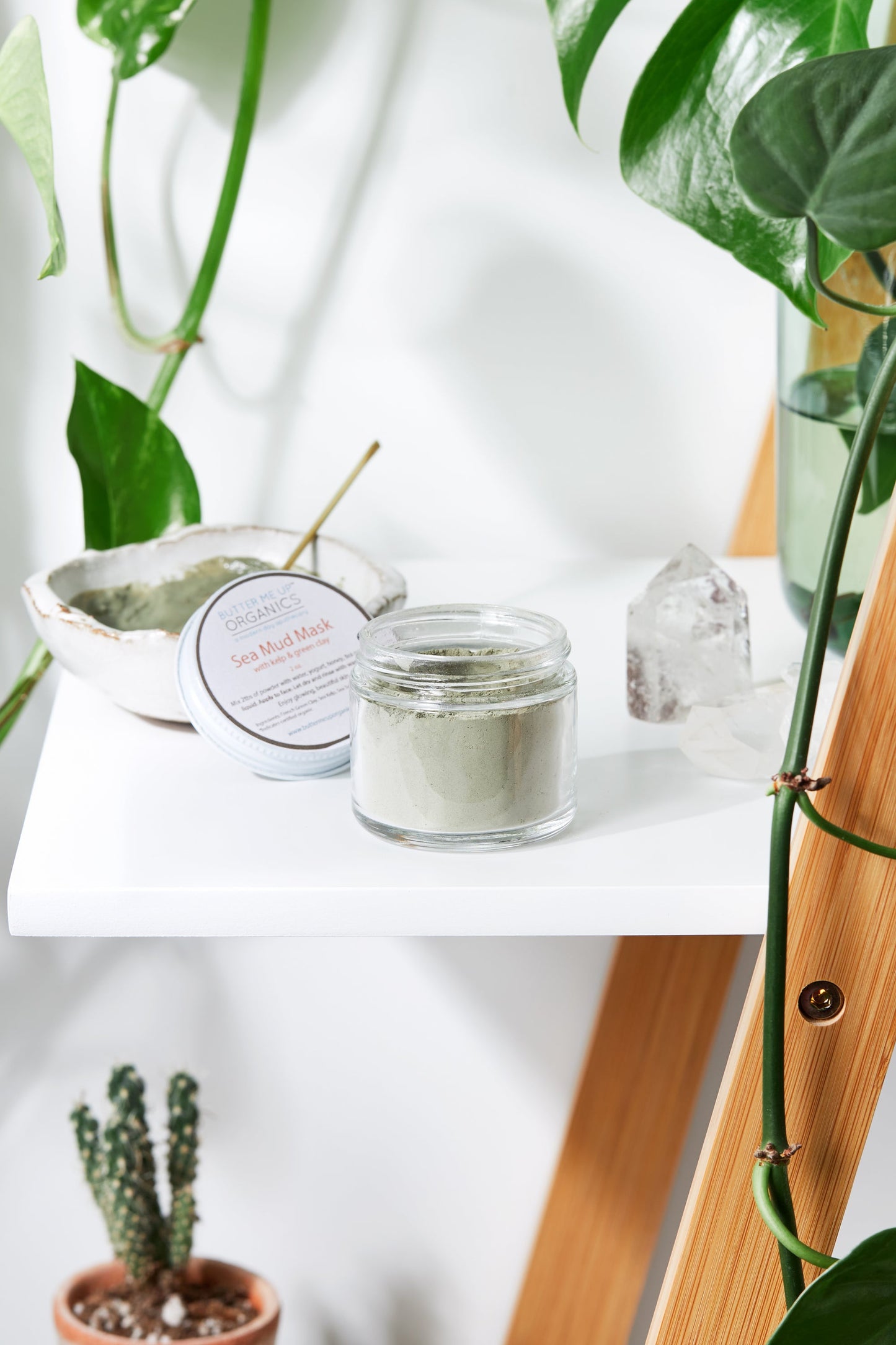 A jar of White Smokey Sea Mud Mask, made with French Green Clay and Kaolin Clay, rests on a white shelf amid lush plants, a crystal, and a brush-filled bowl—ideal for natural skin detoxification.