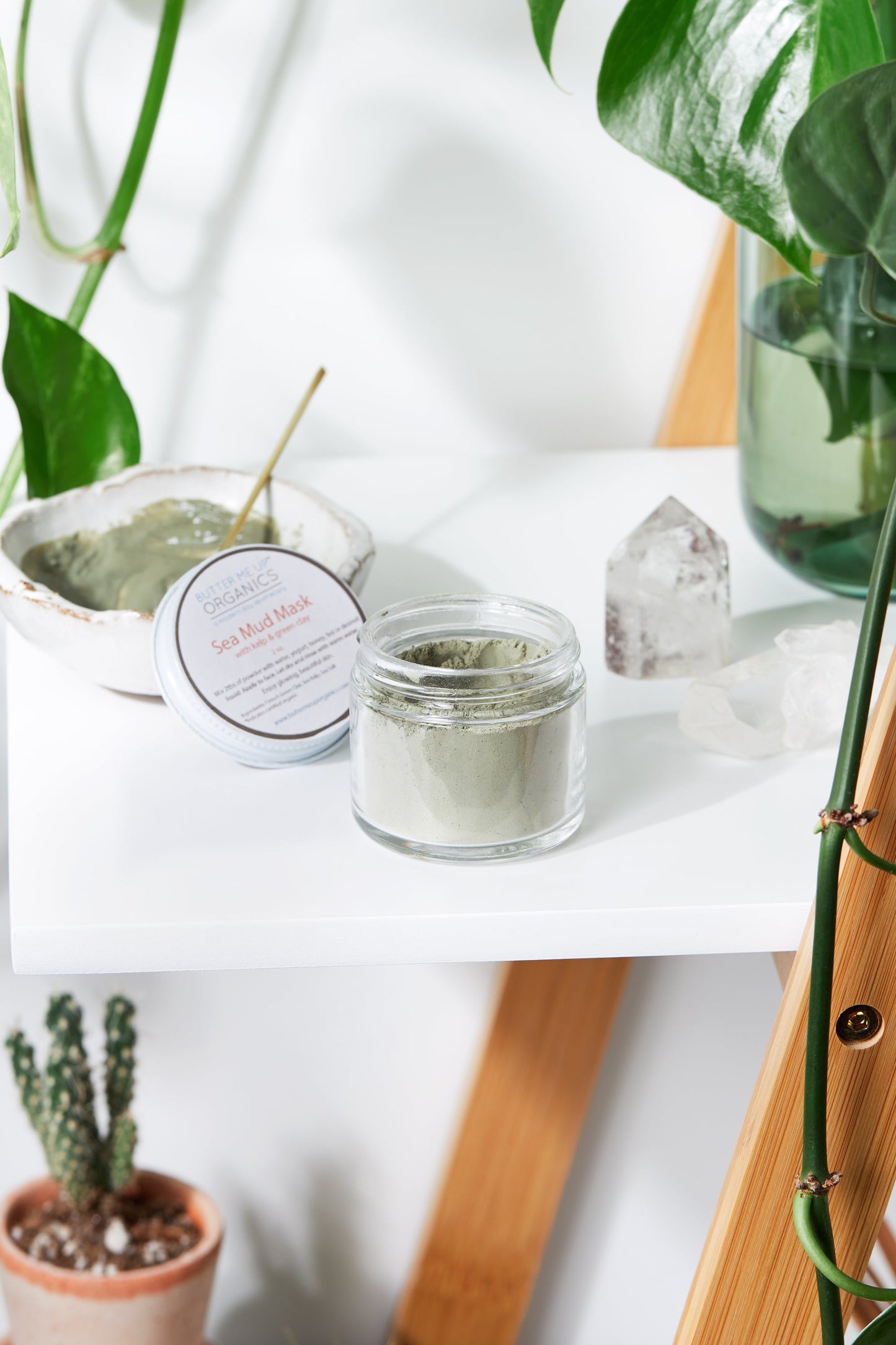 A jar of White Smokey's Sea Mud Mask / French Green Clay Kaolin Clay sits on a white shelf beside a small bowl, a crystal, and potted plants.