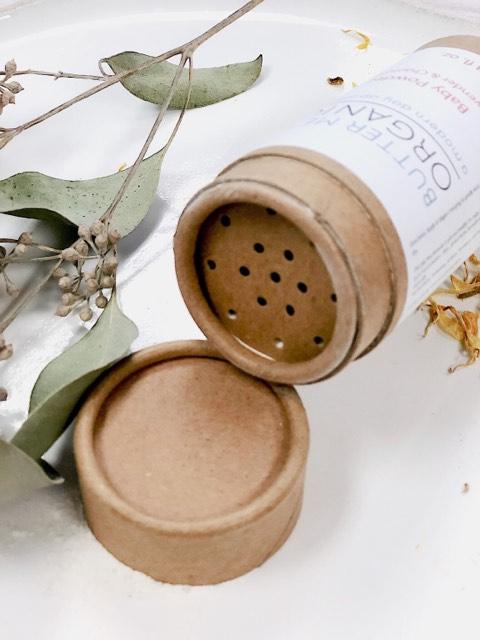 A tilted open shaker with a cork lid, containing White Smokey All Natural Talc Free Baby Powder, surrounded by dried eucalyptus leaves and tiny white flowers on a white surface.
