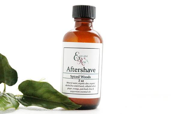 Maroon Oliver's "Spiced Woods Natural Aftershave" in a brown bottle with a white label, rests next to green leaves, offering a refreshing natural aftershave experience.