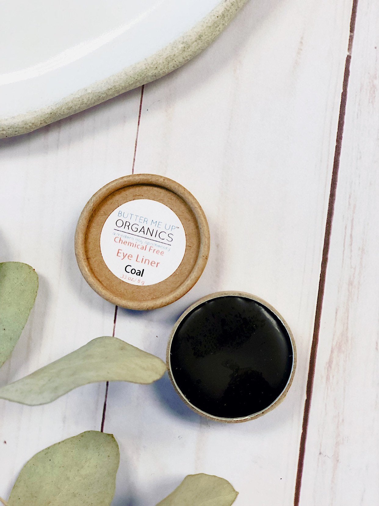 A container of coal-colored eyeliner labeled "White Smokey Organic Eye Coal" sits open on a light wooden surface, highlighting its eco-friendly packaging beside greenery and a plate.