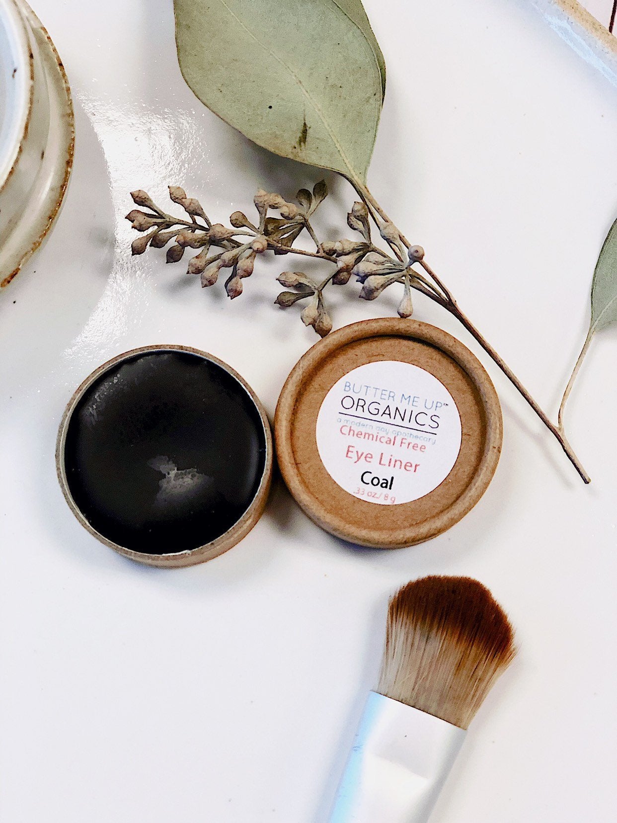 An open container of White Smokey's Butter Me Up Organic Eyeliner rests on a white surface with a makeup brush and dried leaves. Made from organic ingredients, it boasts eco-friendly packaging, offering a mindful beauty choice.