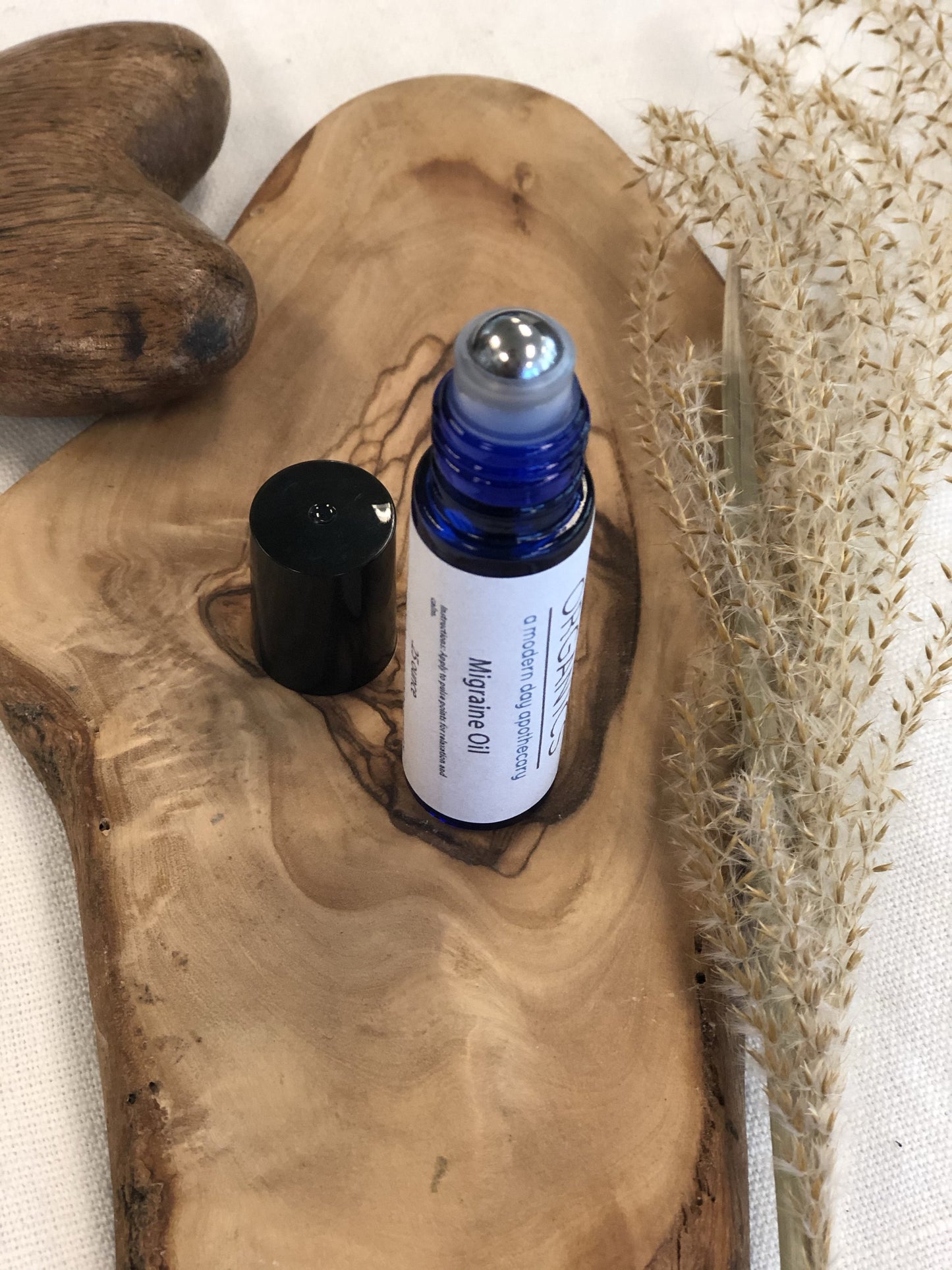 The White Smokey Essential Oil for Migraines, in a blue roller bottle with a black cap, is displayed on a wooden board with dried grasses and a small wooden heart.