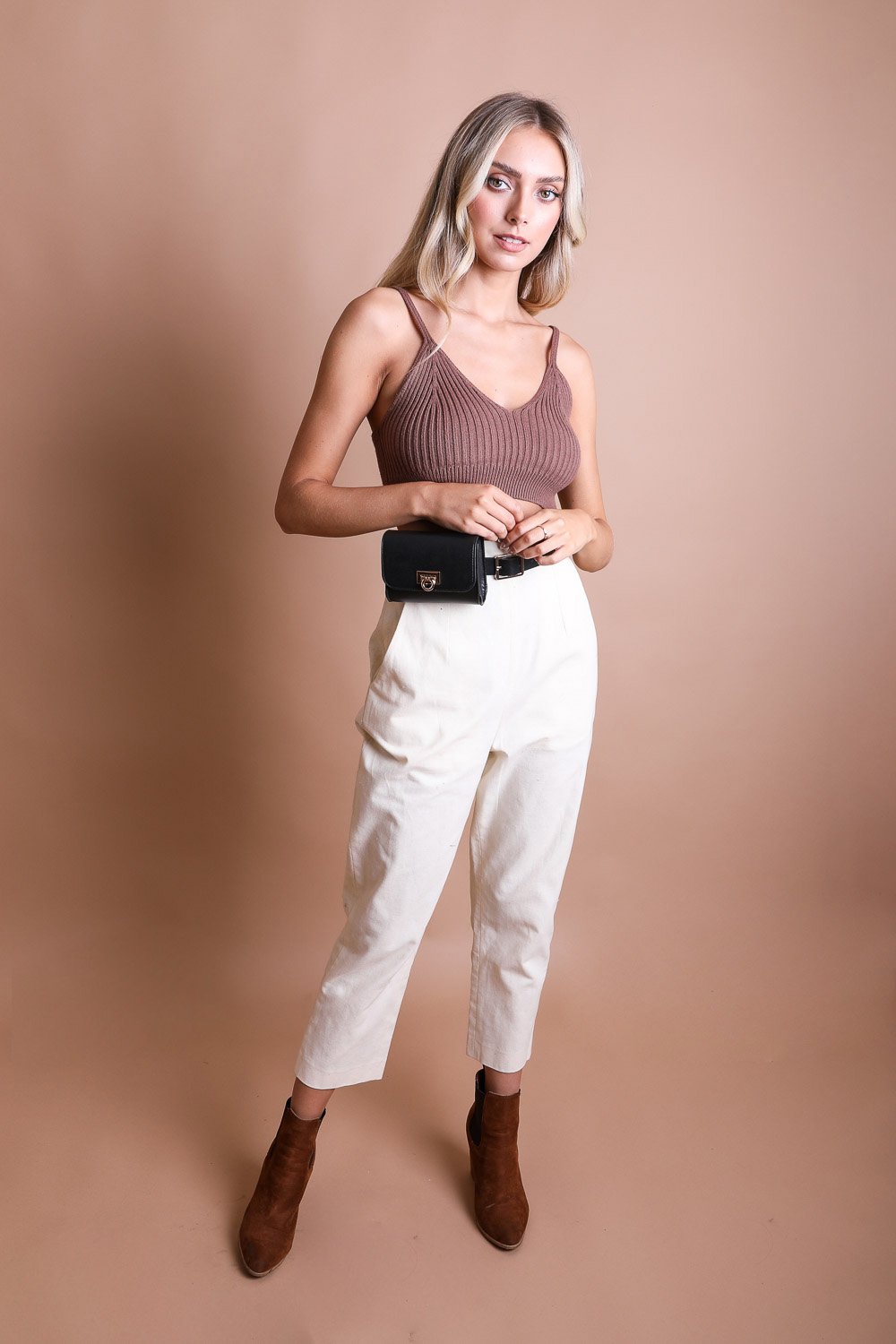 A woman stands against a beige background, wearing a brown sleeveless top, white high-waisted pants, brown ankle boots, and a chic Periwinkle Aether Versatile Vegan Leather Belt Bag.