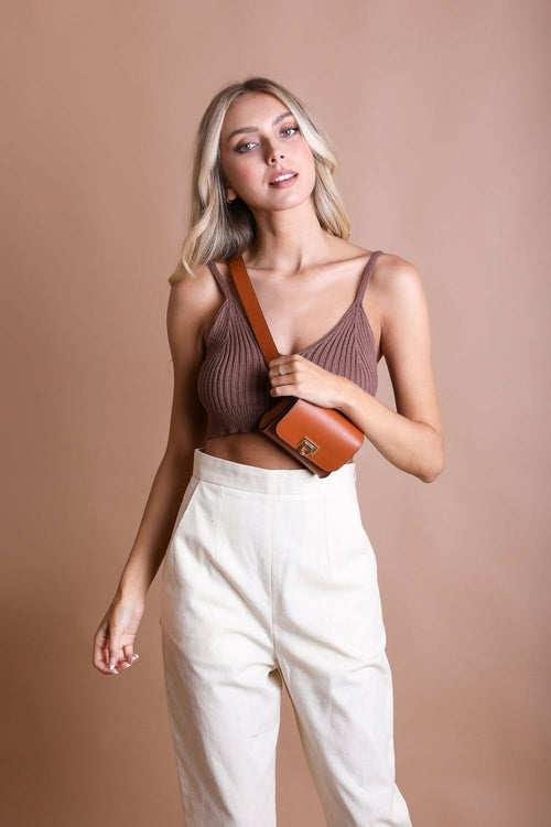 A woman in a brown crop top and high-waisted white pants poses against a plain beige background, holding the strap of a chic Periwinkle Aether Versatile Vegan Leather Belt Bag.