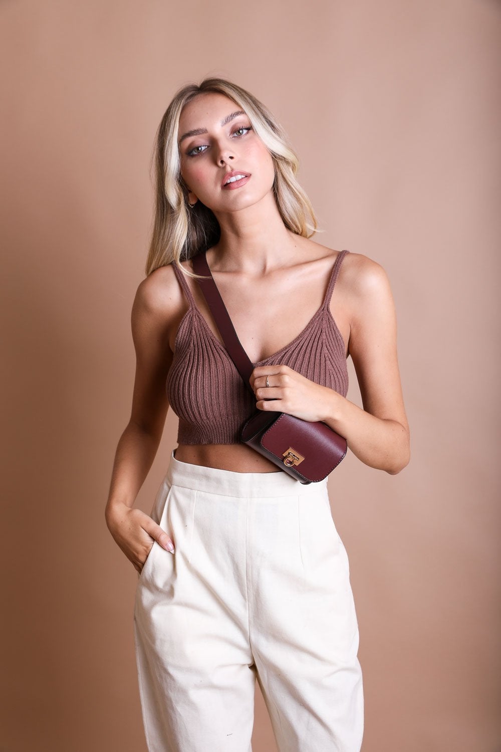 A woman with long blonde hair and a neutral expression wears a brown crop top with white high-waisted trousers, complemented by a chic accessory: the Versatile Vegan Leather Belt Bag in maroon from Periwinkle Aether across her chest, standing against a beige background.