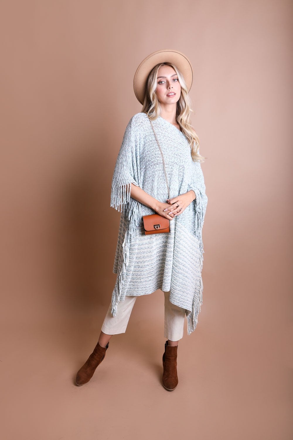 A person wearing a beige hat, gray fringed poncho, white pants, and brown boots stands against a plain beige background, holding the Periwinkle Aether Versatile Vegan Leather Belt Bag—a chic accessory that adds a touch of style to the ensemble.
