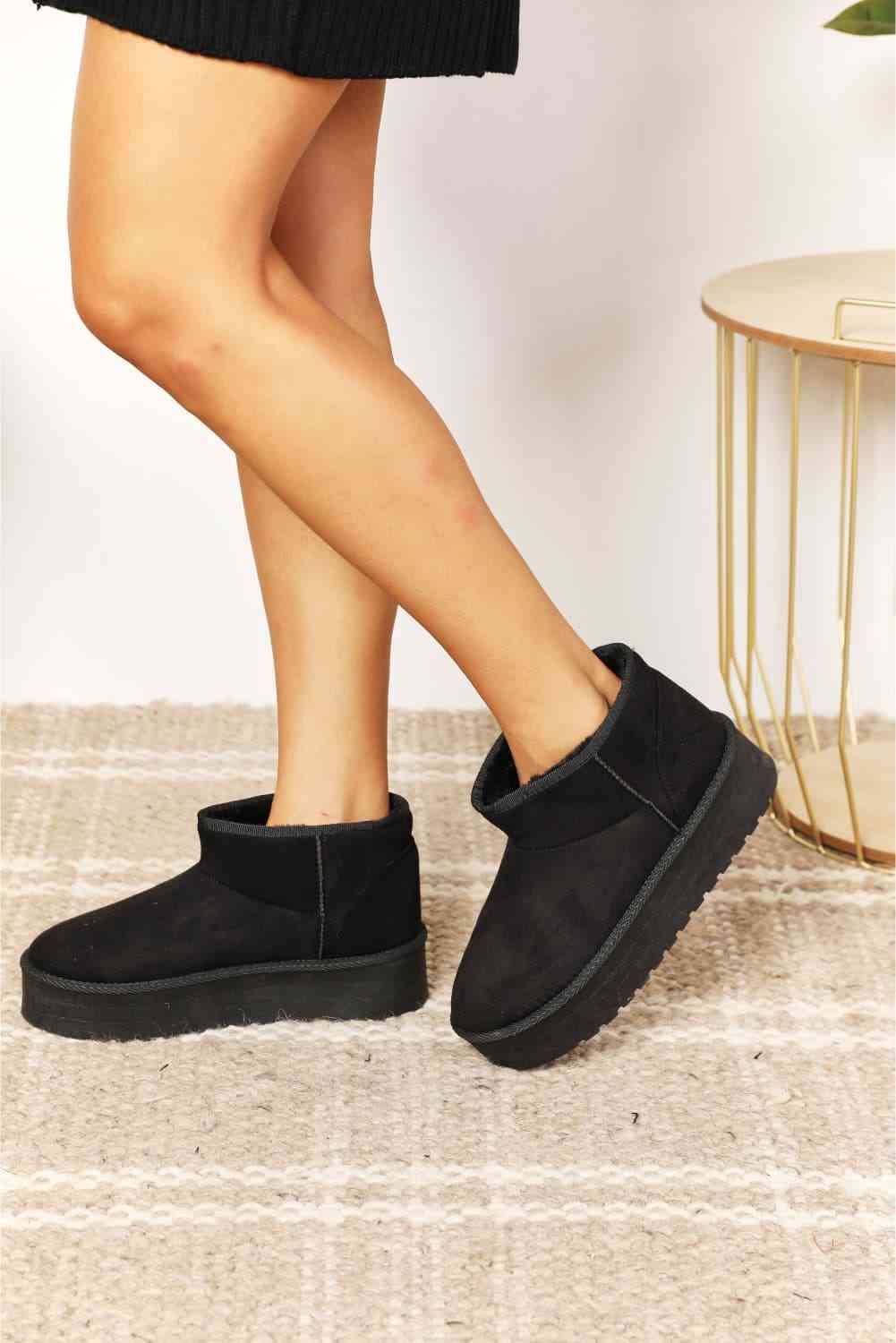 A person wearing Legend Women's Fleece Lined Chunky Platform Mini Boots by Trendsi stands on a light-colored textured carpet next to a small round table.