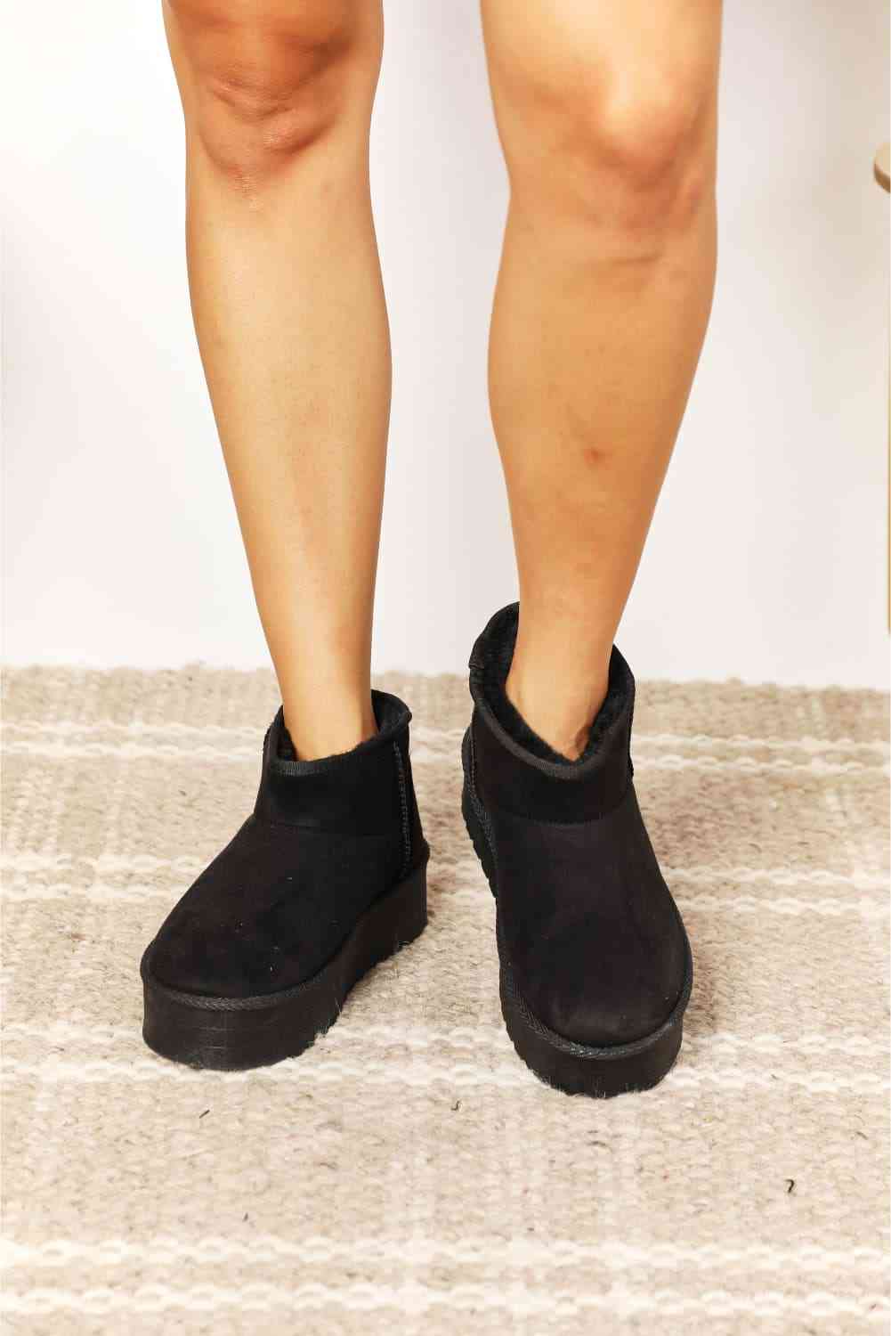 A person's lower legs are shown wearing the Legend Women's Fleece Lined Chunky Platform Mini Boots by Trendsi, in black, on a patterned rug.