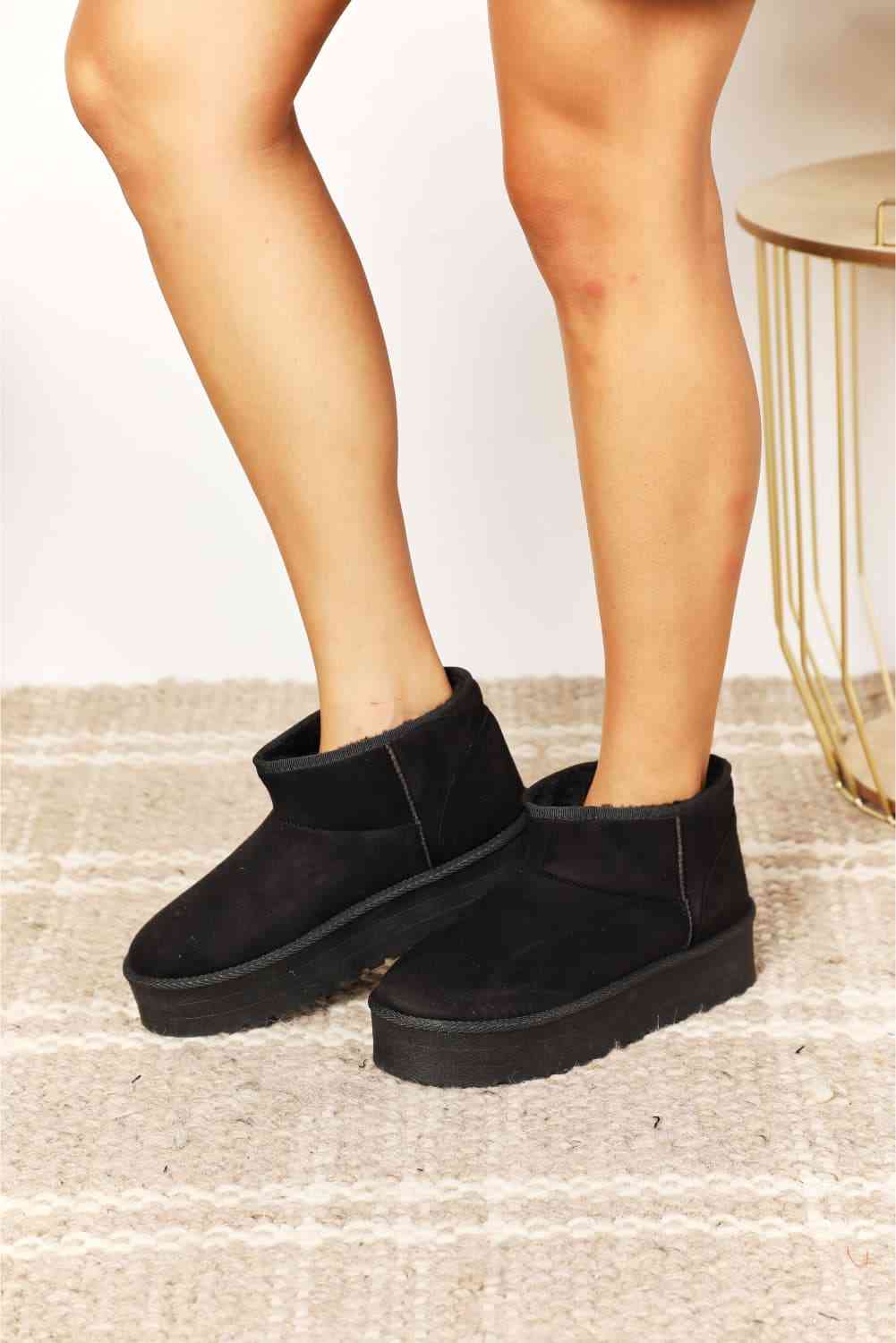 A person standing on a light-colored rug, wearing black Trendsi Legend Women's Fleece Lined Chunky Platform Mini Boots.