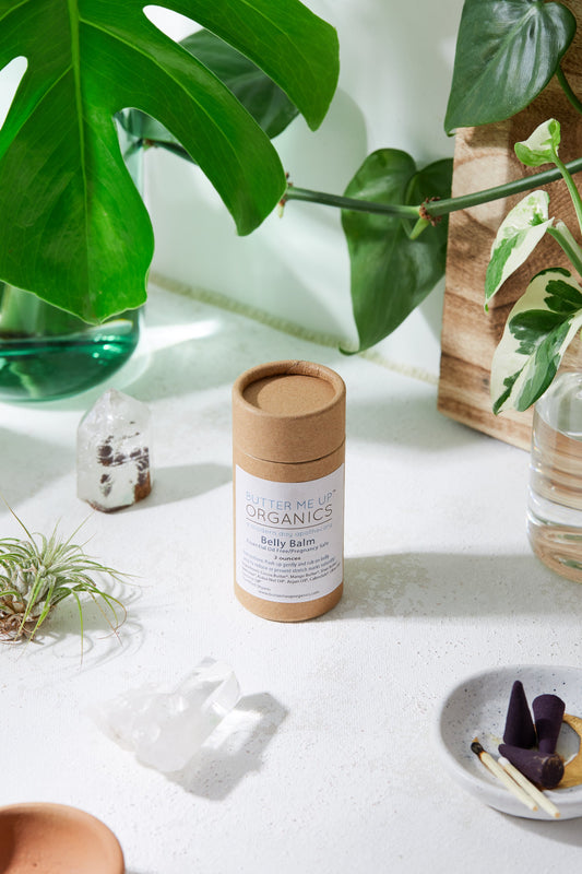 A cylindrical butter balm container labeled "White Smokey - Belly Balm / Organic Pregnancy Balm / Stretch Mark Balm" is positioned on a white surface among plants, crystals, and a small dish with matchsticks. This hydrating skin care product aims for effective stretch mark prevention.