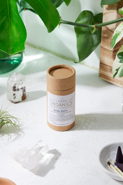 A cardboard tube labeled "White Smokey Belly Balm / Organic Pregnancy Balm / Stretch Mark Balm" is placed on a white surface surrounded by green plants and crystals, highlighting its hydrating skin care and stretch mark prevention properties.