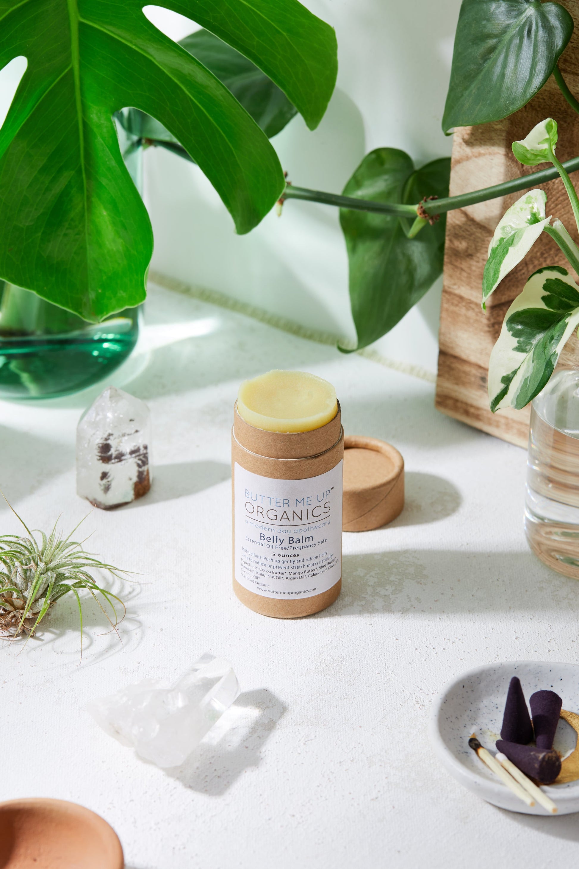 A container of Belly Balm from White Smokey sits on a white surface, surrounded by green plants and small decorative crystals, poised to offer hydrating skin care and stretch mark prevention.