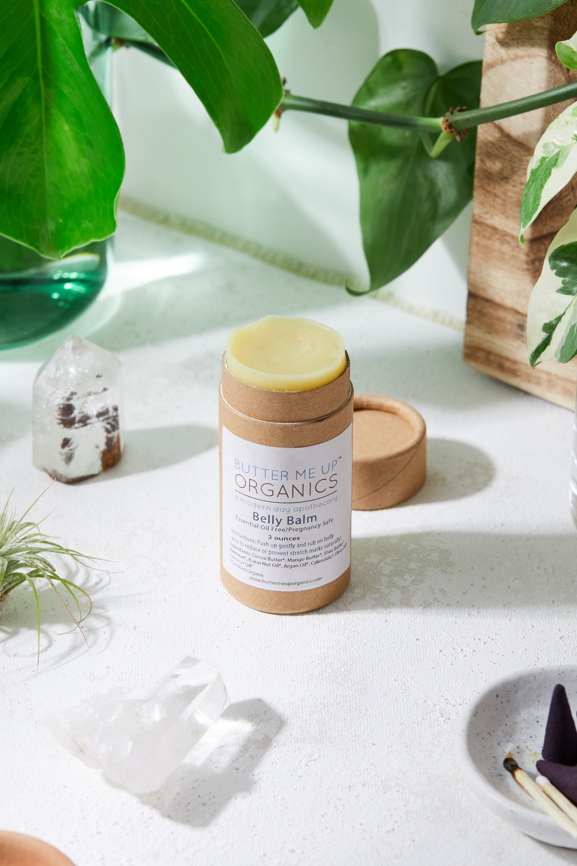 A cylindrical container of "White Smokey Belly Balm / Organic Pregnancy Balm / Stretch Mark Balm" placed on a white surface, surrounded by plants and crystals, showcases its commitment to hydrating skin care and stretch mark prevention.