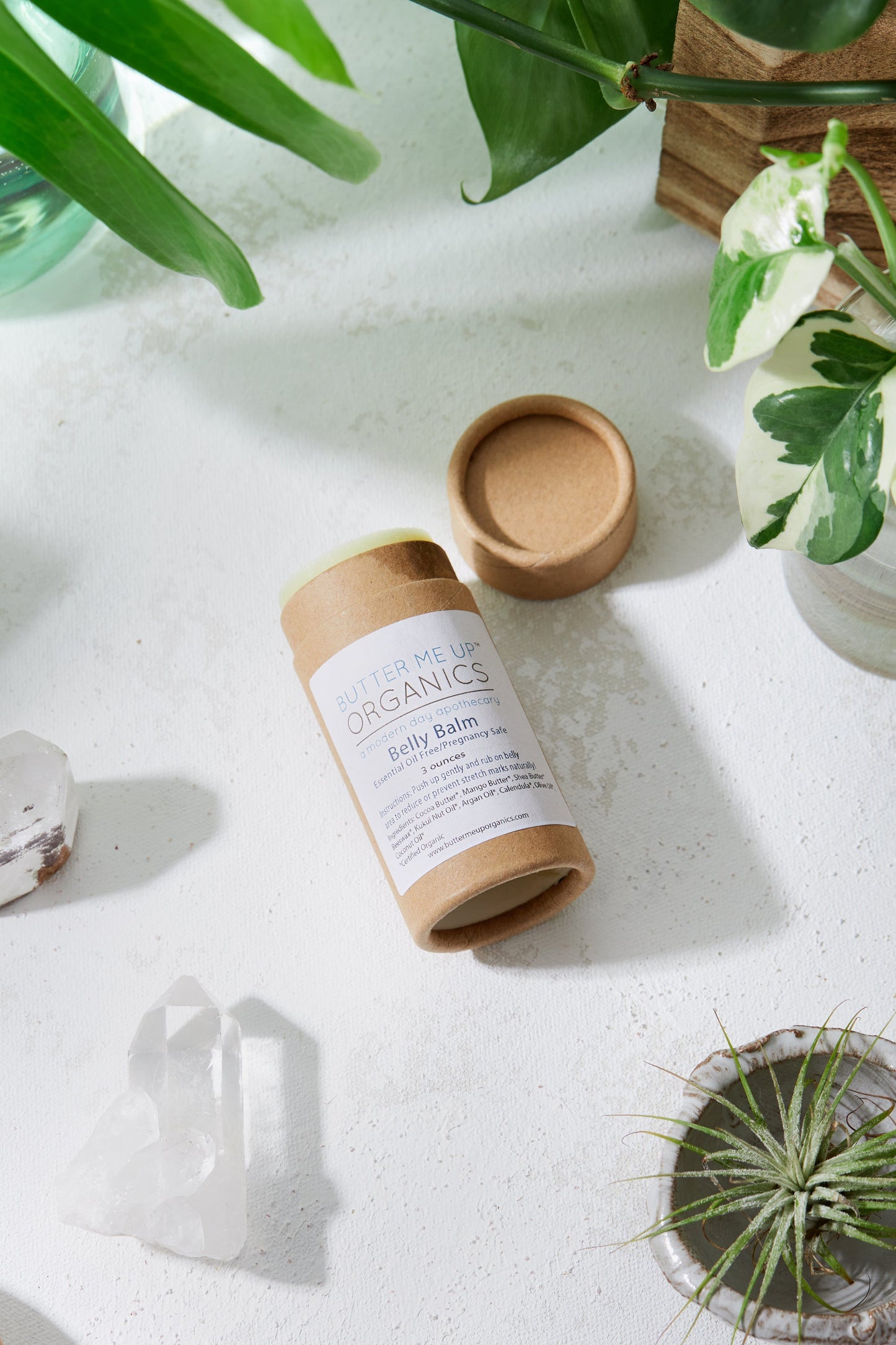 A biodegradable cardboard tube labeled "White Smokey Belly Balm" lies opened on a white surface, surrounded by plants and crystals, offering a blend of organic belly balm for stretch mark prevention and hydrating skin care.