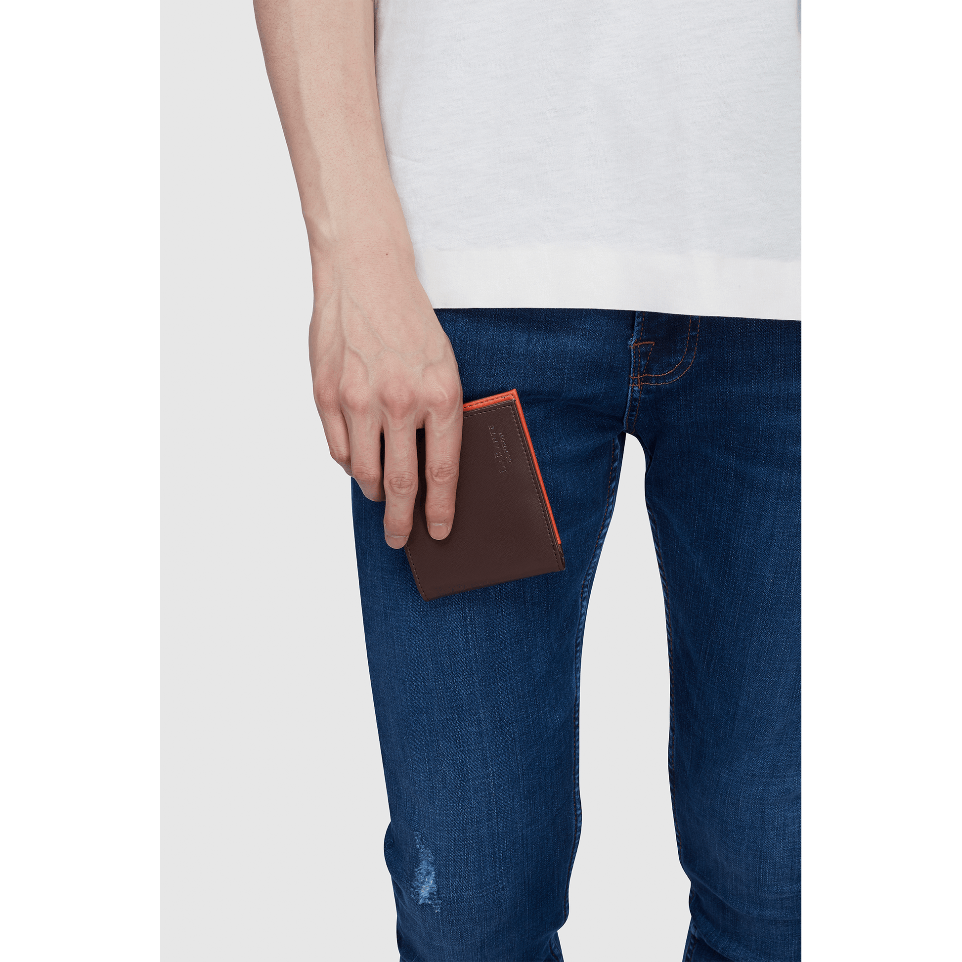 Person wearing a white shirt and blue jeans, holding a Brown - Brave Vegan Bifold Wallet by Jade Azolla with an orange edge near their pocket.