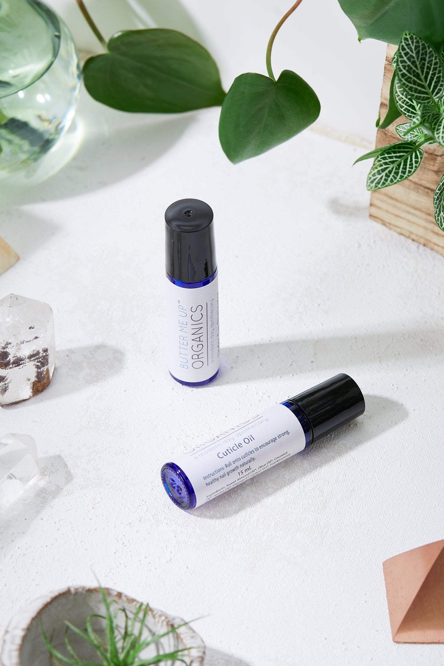 Two bottles of White Smokey Organic Cuticle Oil arranged on a table with green plants and decorative stones, illuminated by natural light, containing organic ingredients.