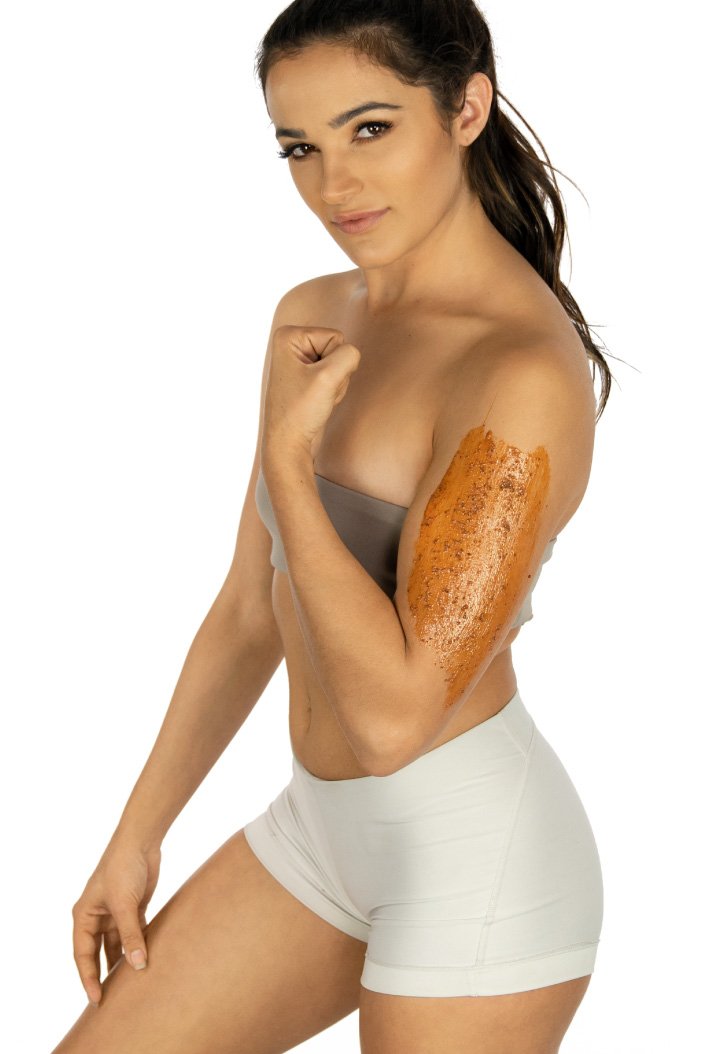 Woman in fitness attire shows her flexed arm with a brown Cyan Ares Brightening Exfoliating Mask applied, posing against a white background.