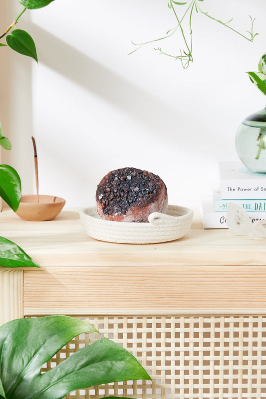 A brown and black Organic Geode Bath Bomb from White Smokey sits in a woven tray on a wooden surface, surrounded by green plants, books, and a Himalayan salt lamp.