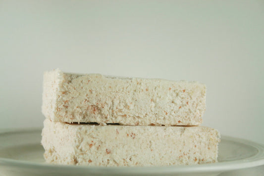 Two rectangular pieces of a crumbly, beige dessert are stacked on a white plate against a plain background, resembling the calm and natural aesthetic of Maroon Oliver's Himalayan Salt Soap - Babassu Salt Bar - Essential.