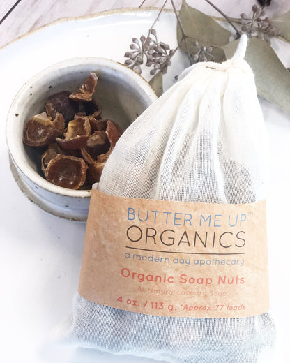 A bag labeled "White Smokey Organic Soap Nuts / All Natural Laundry Soap / Eco friendly" sits next to a bowl with soap nuts on a white surface, adorned with decorative leaves, renowned for their natural surfactant properties.