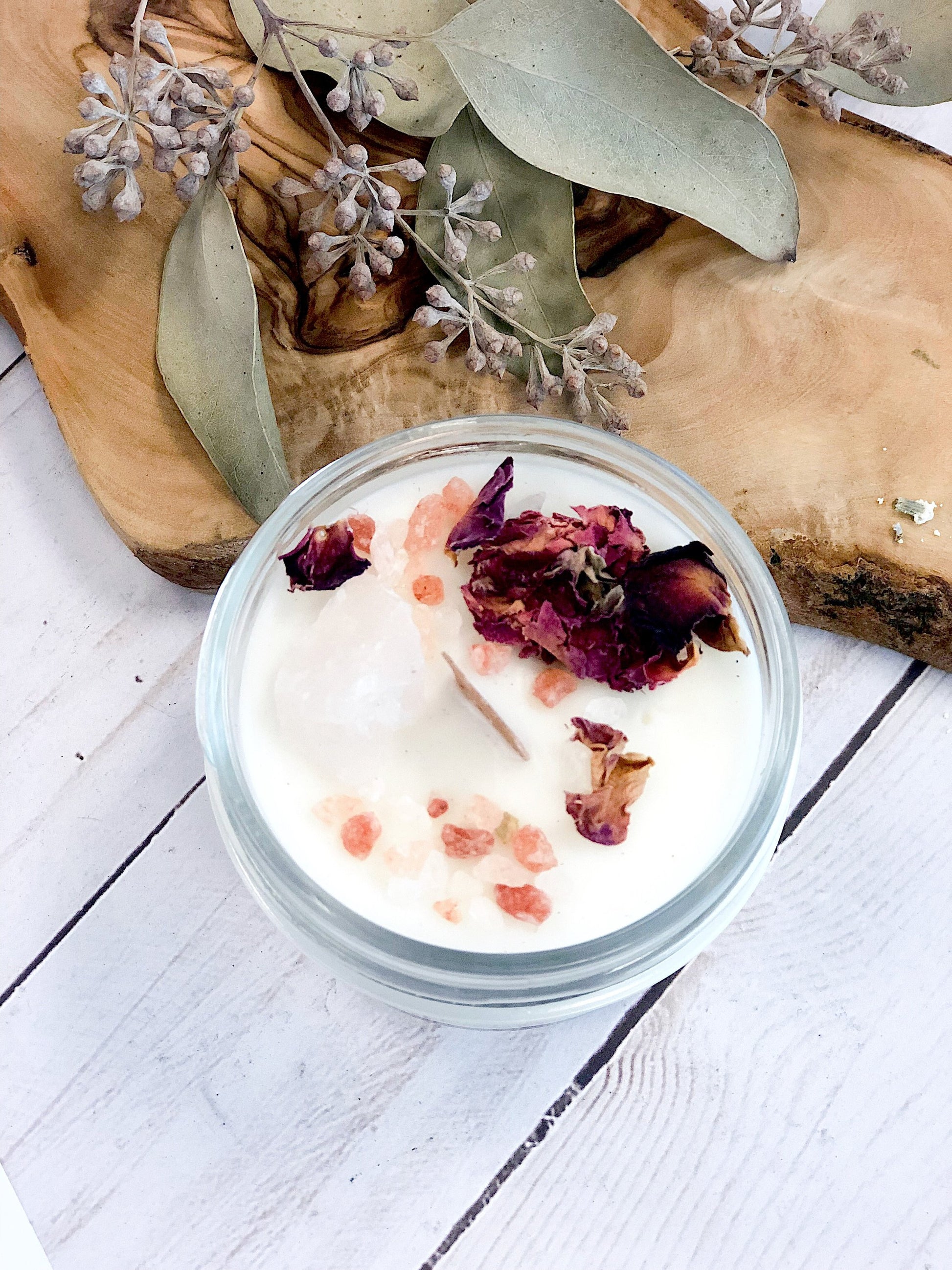 Sentence with replacement: A jar of Love Spell Intention Candle with rose quartz crystals and dried flowers on top, placed on a wooden surface next to dried plants by White Smokey.