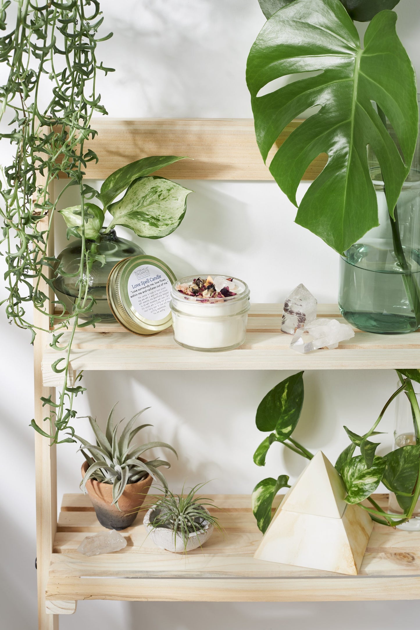 A wooden shelf displaying assorted plants, candles, a rose quartz crystal, and geometric decor in a brightly lit room with a White Smokey Love Spell candle.