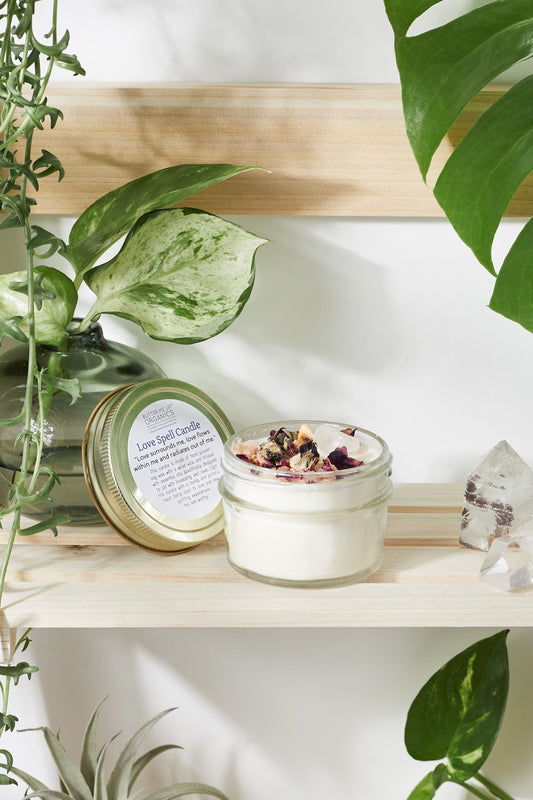 A jar of Love Spell body cream by White Smokey on a wooden shelf beside a metal tin, surrounded by green plants and a rose quartz crystal.