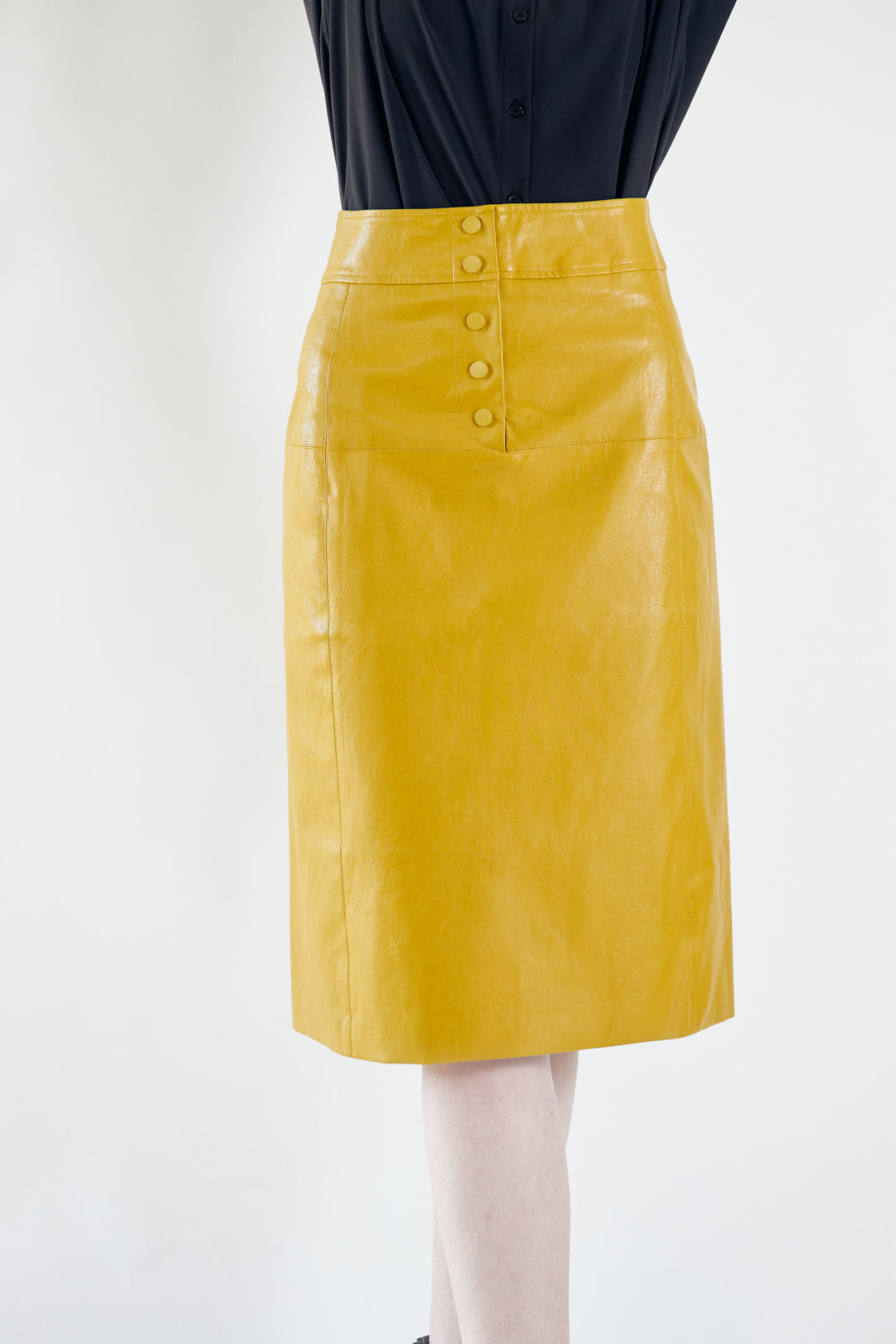 A person standing in a Glossy Vegan Leather Pencil Skirt in mustard yellow with button detailing at the back by Mauve Daisy.