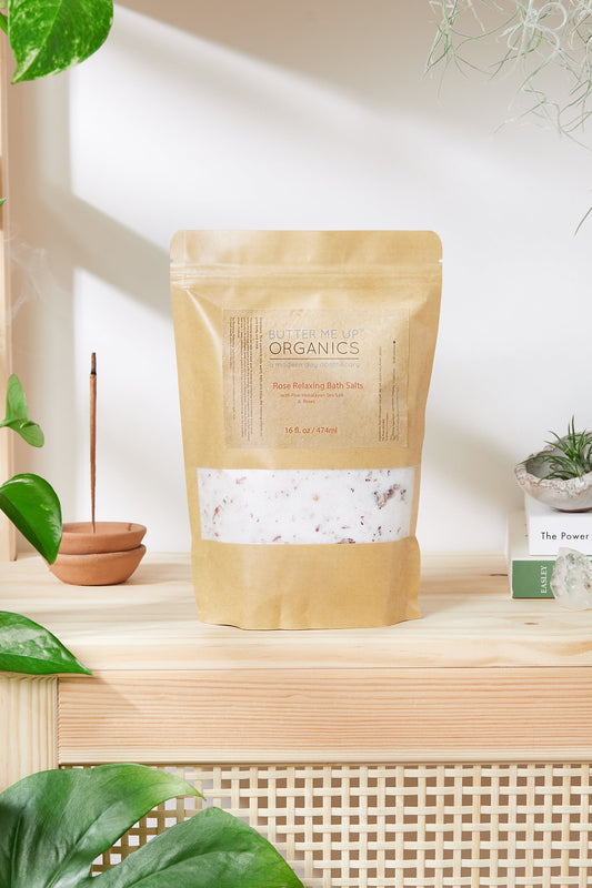 A kraft bag labeled "White Smokey Organic Rose Salt Soak / Epsom Salt Soak / Pink Himalayan Sea Salt" is placed on a wooden surface with plants, books, and an incense holder nearby. Infused with detoxifying salts, this blend promises a rejuvenating experience.