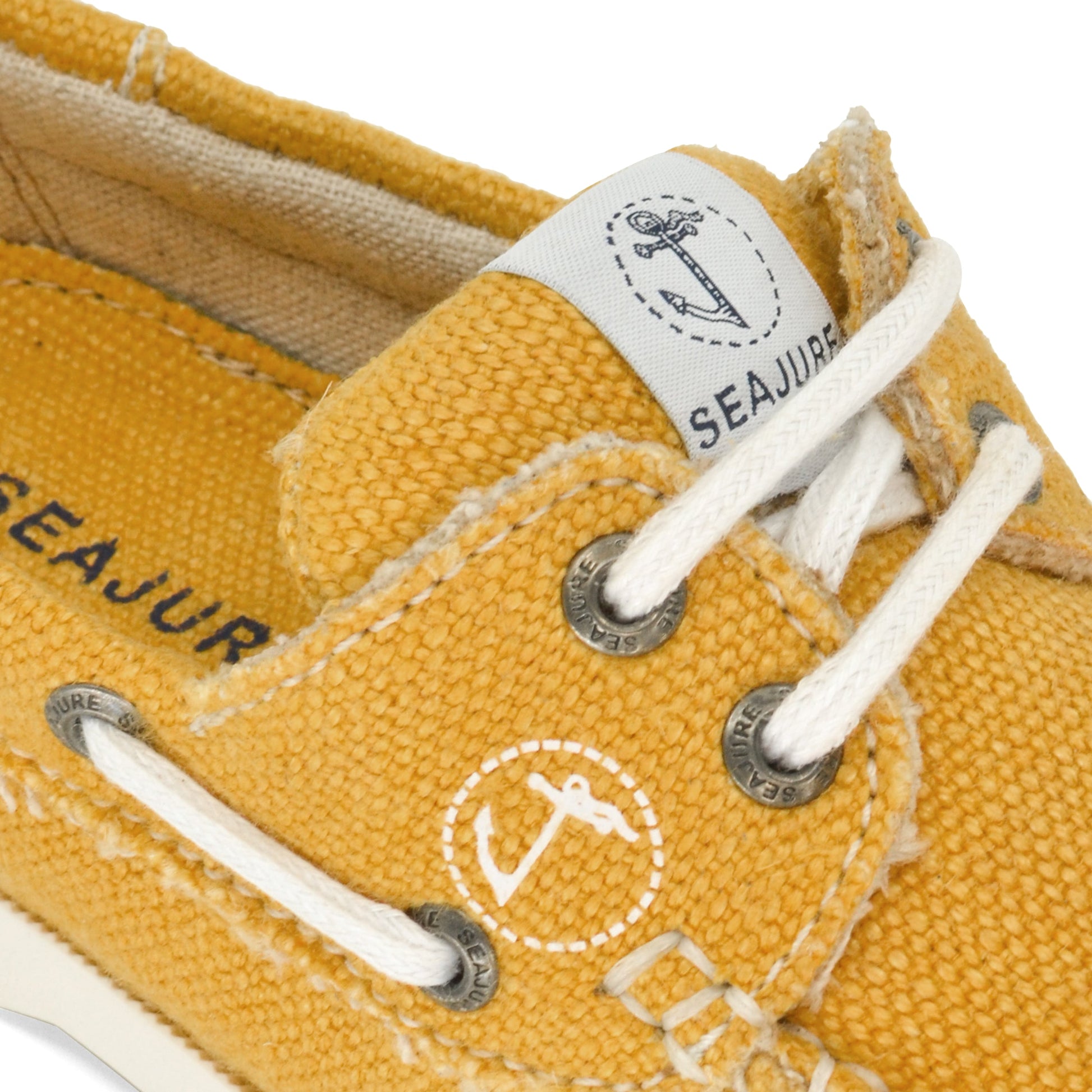 Close-up of a yellow canvas boat shoe with white laces. The shoe features a label with an anchor and the word "AMETHYST HESTIA" along with stitching and metal eyelets, embodying sustainability as part of the Amethyst Hestia Women Hemp & Vegan Boat Shoe Saharun collection.