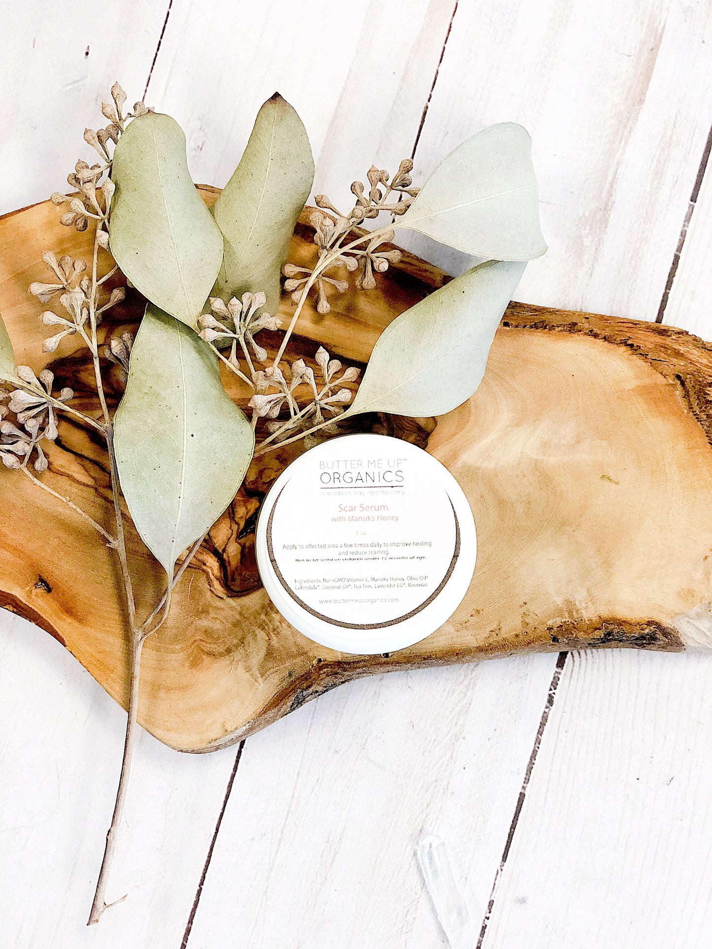 A round container labeled "White Smokey Scar Serum" placed on a wooden tray with dried leaves and a white background, featuring organic skincare ingredients like Manuka Honey for an added boost.