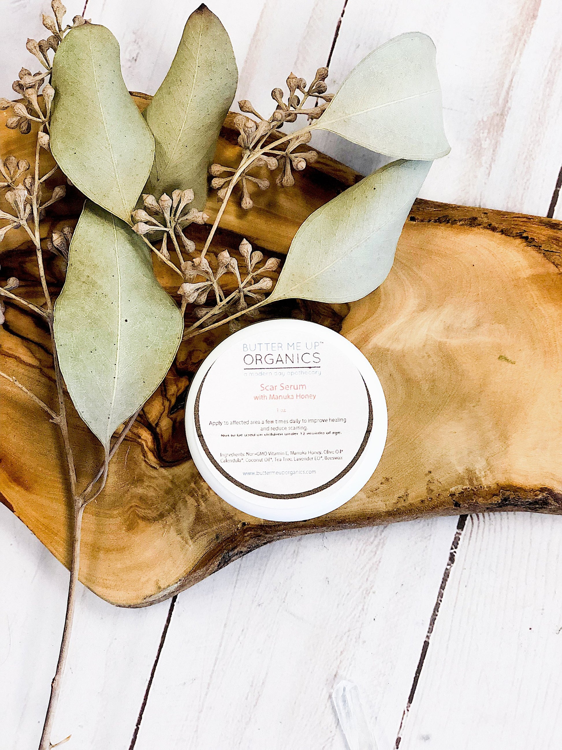 A small container of "White Smokey" Scar Serum sits on a wooden board, surrounded by dry eucalyptus leaves. The label mentions ingredients and usage instructions, highlighting its place in organic skincare routines.