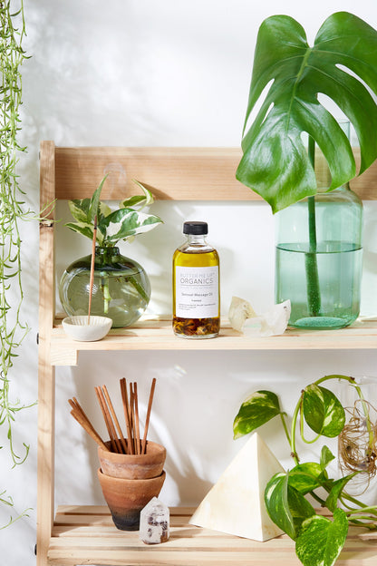 Wooden shelves with various plants in glass containers, a bottle of White Smokey Sensual Massage Oil, terracotta pots, reed diffusers, organic oils, and a pyramid-shaped ornament.