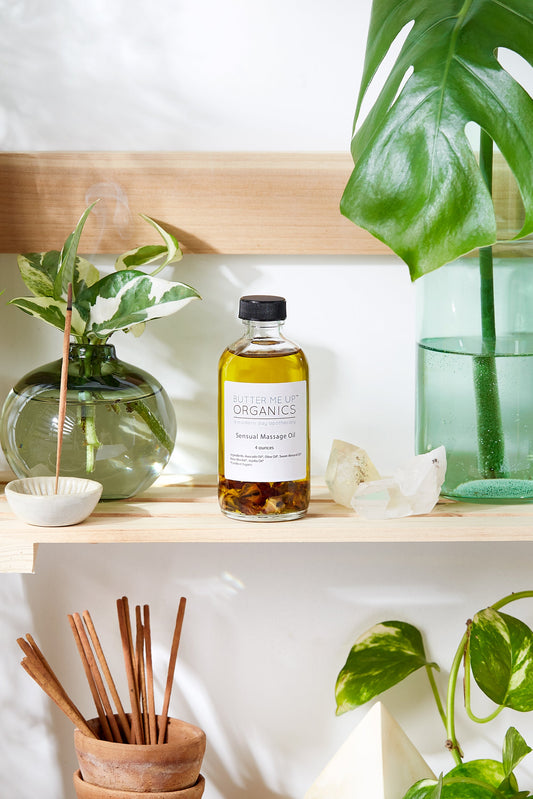 A bottle of Sensual Massage Oil with the label "White Smokey" is placed on a wooden shelf, surrounded by plants and decorative items including incense sticks, crystals, and a delicate hint of Rose Absolut.