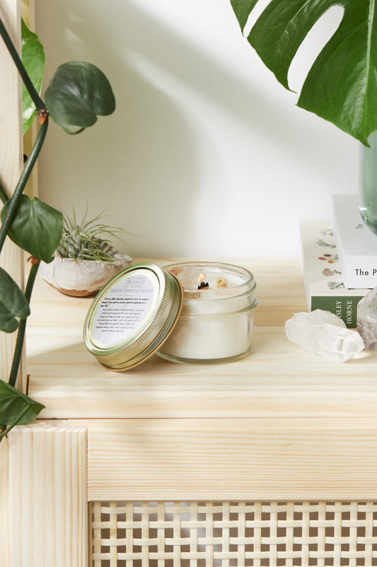 A lit Space Clearing Candle by White Smokey in a glass jar with a gold lid rests on a wooden surface, surrounded by green plants, a small crystal for negative energy cleansing, and a stack of books.