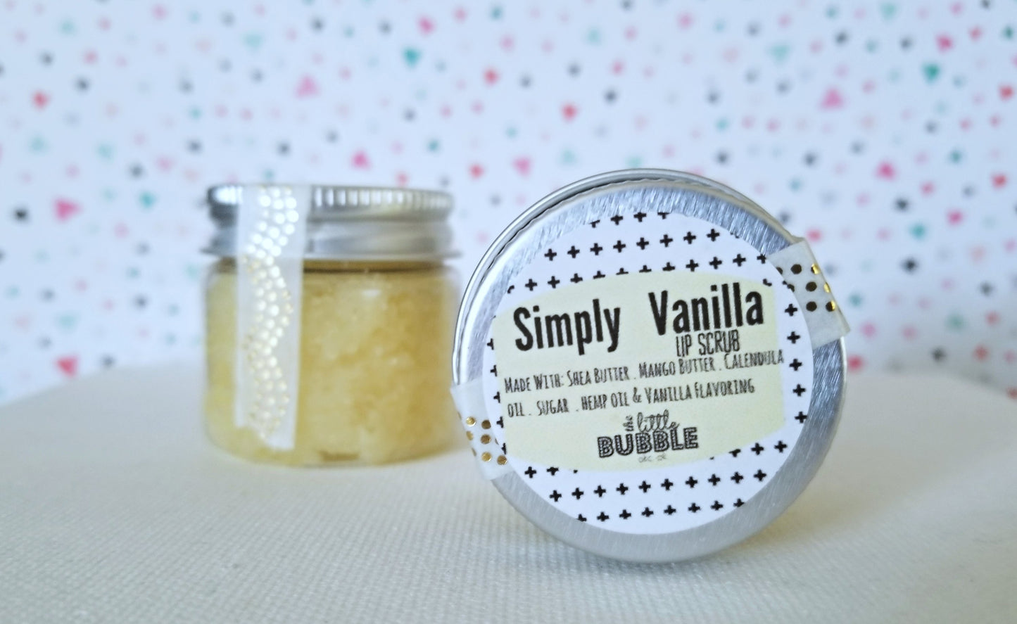 A small jar labeled "Vanilla Lip Scrub" by Magenta Hades, boasting 100% VEGAN ingredients, is displayed against a colorful speckled background. The lid is slightly open, revealing the scrub inside. Perfect to exfoliate and moisturize your lips!