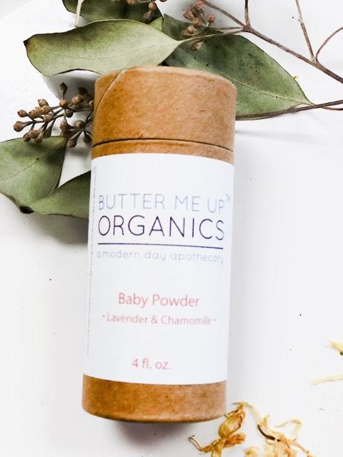 A bottle of "White Smokey" All Natural Talc Free Baby Powder with lavender and chamomile, wrapped in brown paper, surrounded by green leaves and dried flowers on a white background.