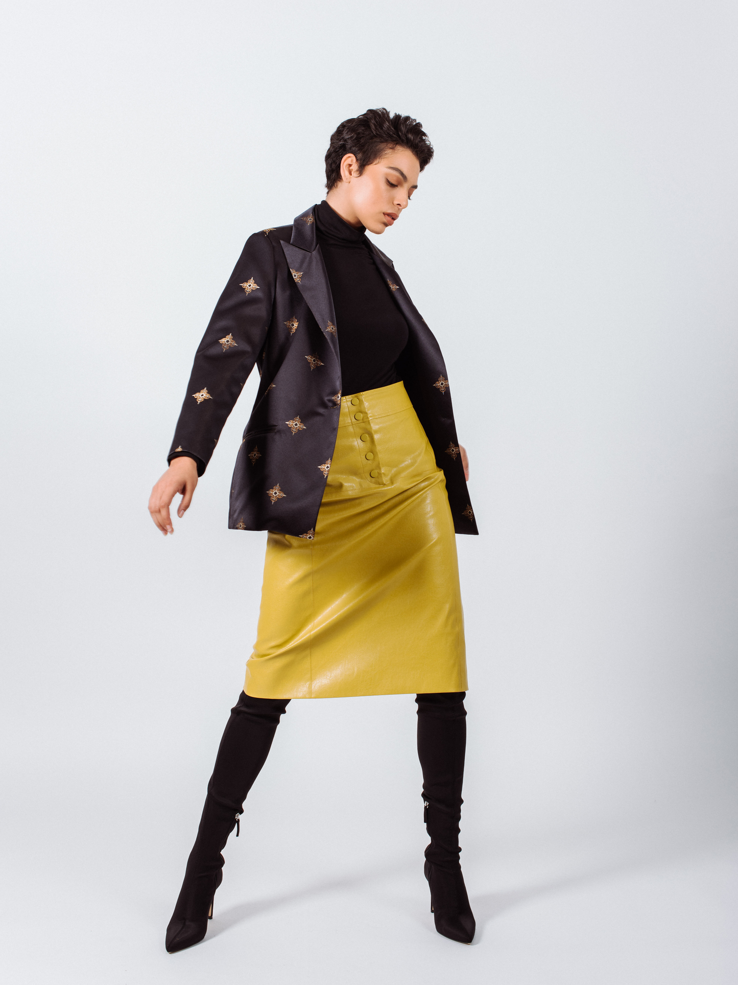 Woman in a black turtleneck, star-patterned jacket, Glossy Vegan Leather pencil skirt from Mauve Daisy, and black boots posing against a plain background.